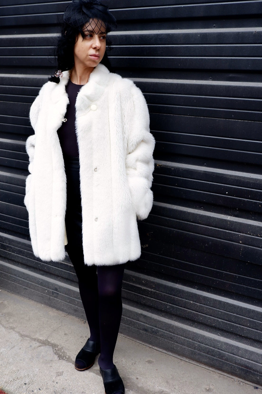 Front view of an ivory faux fur coat worn with black tights against a black ribbed wall