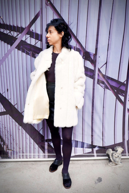 Model in a vintage Monterey ivory faux fur coat and black veiled headband against an industrial mural, coat open to show the satin lining