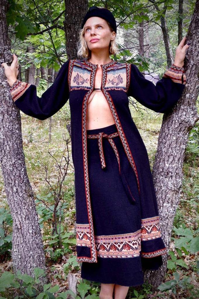 John Ashpool dark knit duster worn open with matching skirt and wrap belt, leaning between trees