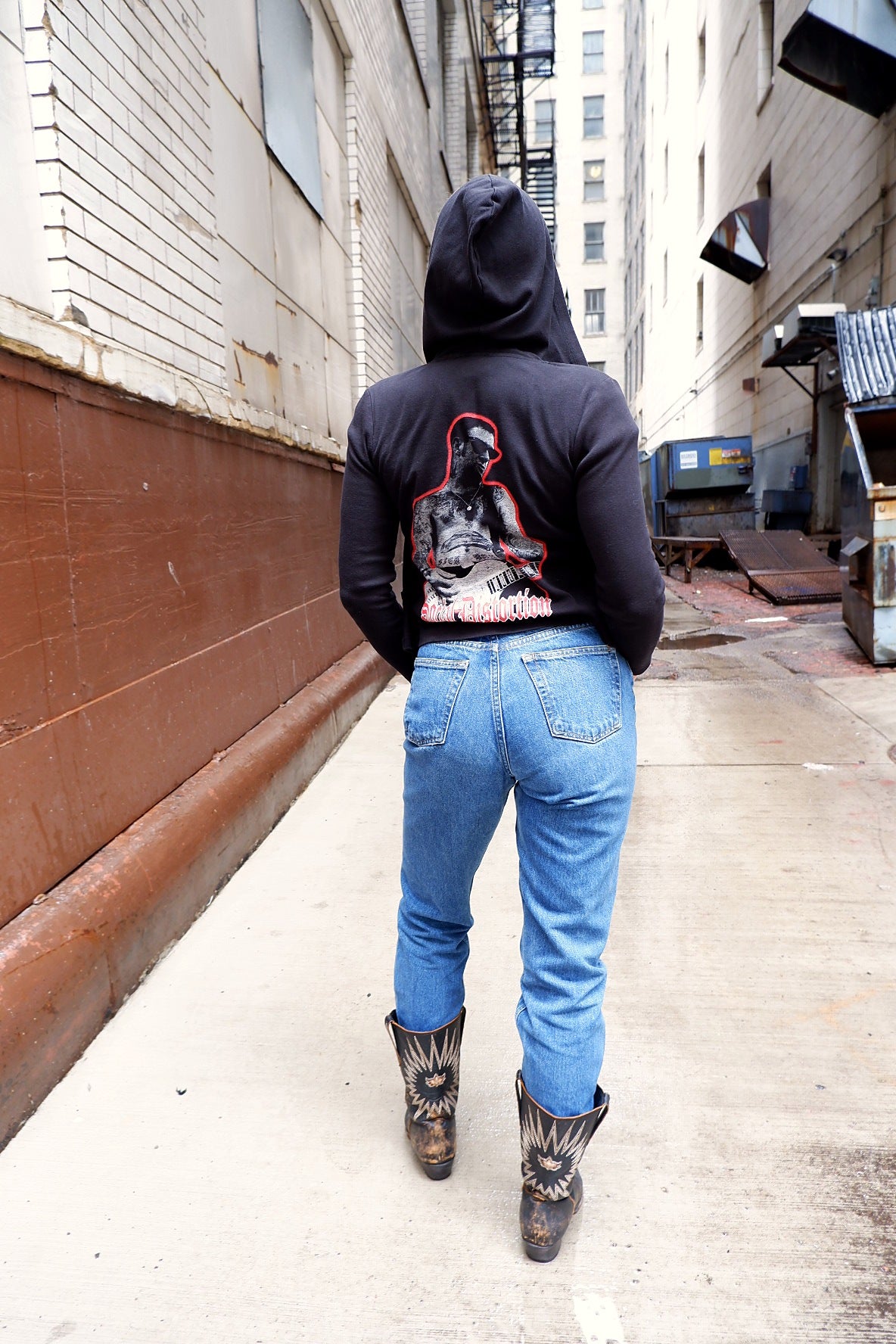 Model walking down a city alley in a black Social Distortion hoodie, jeans and cowboy boots