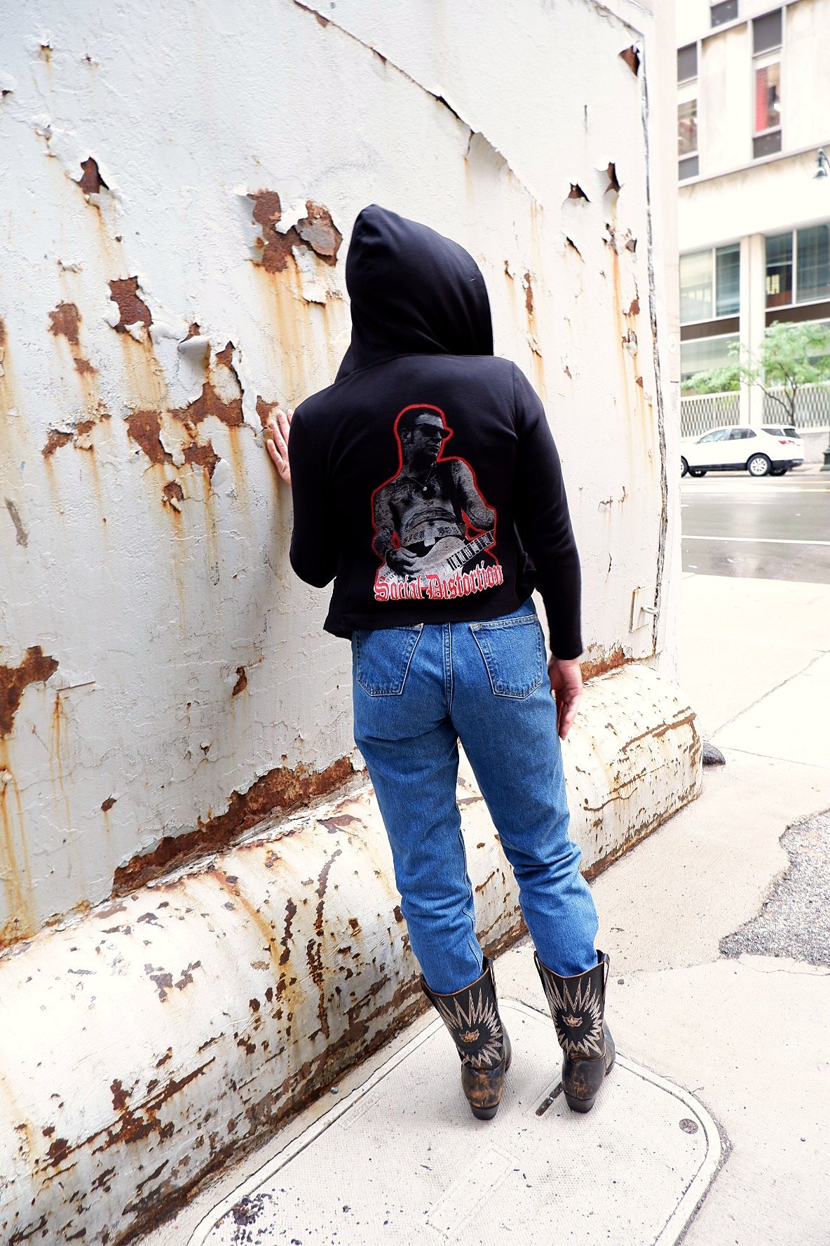 Model in a black Social Distortion hoodie, hood up, leaning on a rusted white wall with vintage jeans and cowboy boots